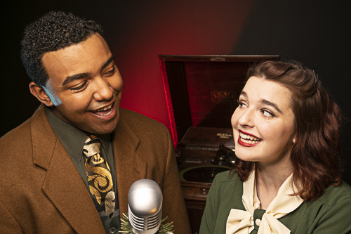 Two actors smile at one another as they sing into a vintage radio microphone.